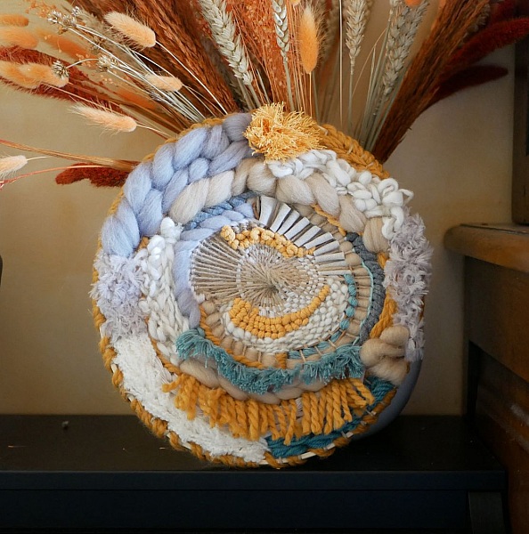 Tissage mural rond contemporain aux textures bohèmes et pompons.