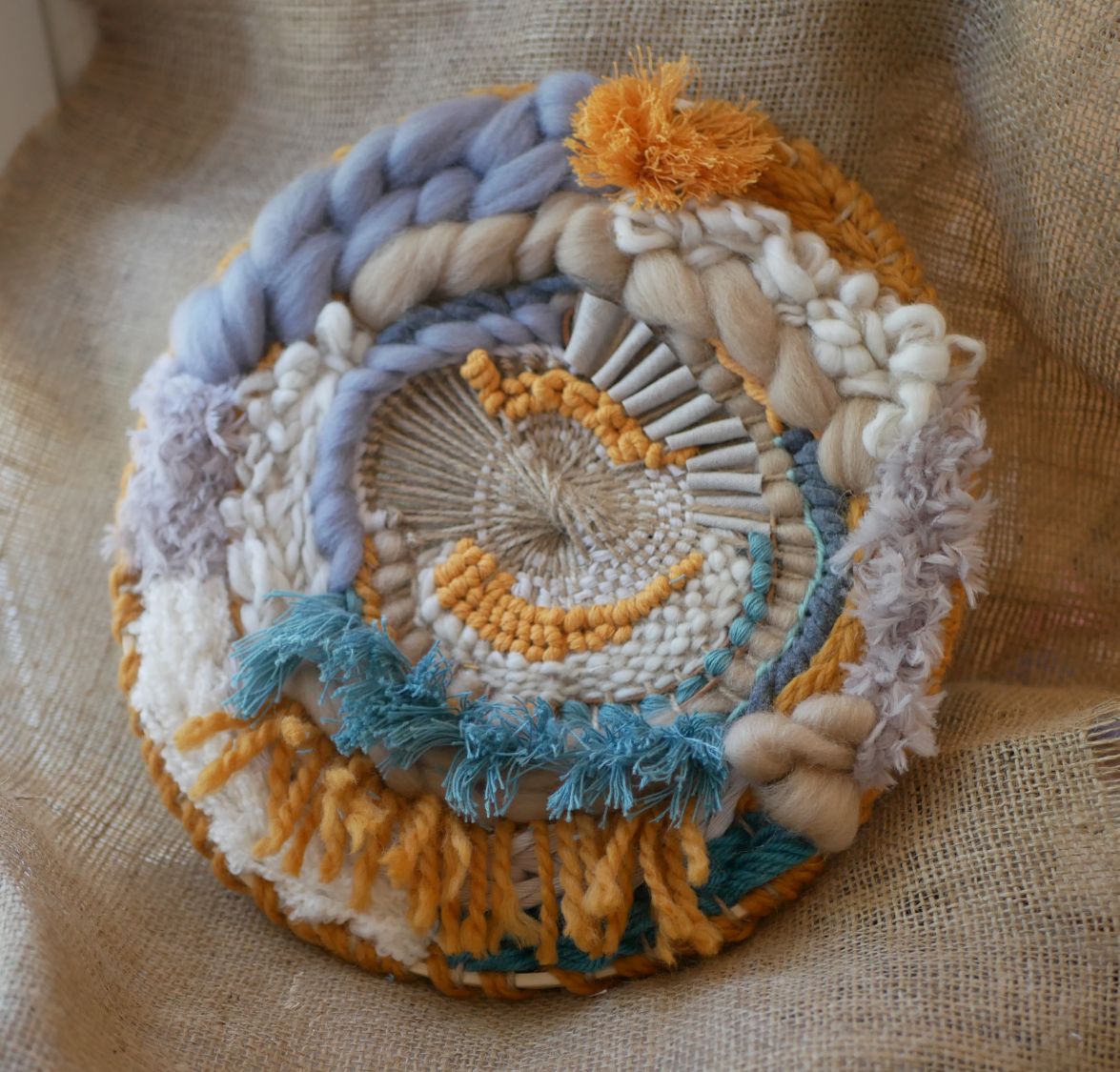 Tissage mural rond contemporain aux textures et pompons bohèmes.