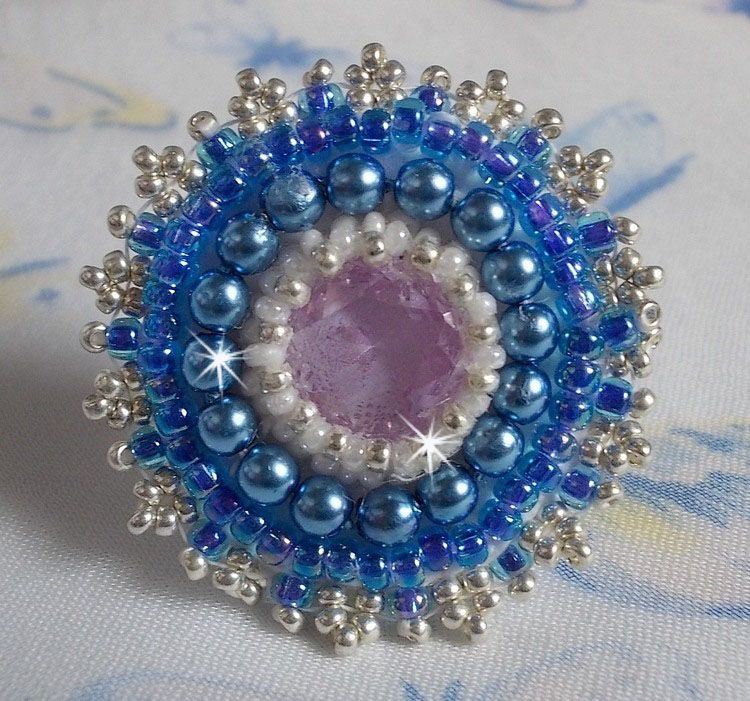 Bague Azur brod&eacute;e avec un cabochon en Cristal de couleur Antique Pink &agrave; laquelle s'ajoute des rocailles et perles rondes nacr&eacute;es. Le tout est mont&eacute; sur une bague filigran&eacute;e Blanche