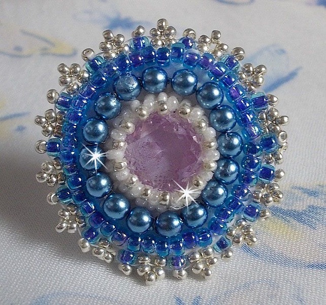 Bague Azur brod&eacute;e avec un cabochon en Cristal de couleur Antique Pink &agrave; laquelle s'ajoute des rocailles et perles rondes nacr&eacute;es. Le tout est mont&eacute; sur une bague filigran&eacute;e Blanche