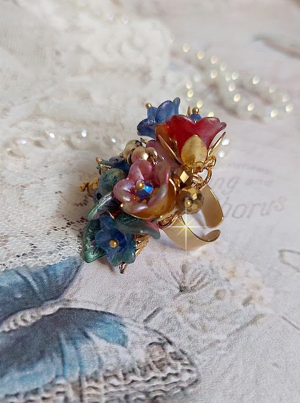 Bague Bel Air mont&eacute;e avec des fleurs lucites peintes &agrave; la main aux couleurs : Bleu, Rose, Rouge, Argent, Blanc avec feuilles ondul&eacute;es Vert Cuivr&eacute; &agrave; laquelle s&rsquo;ajoute perles Dor&eacute;es en Verre