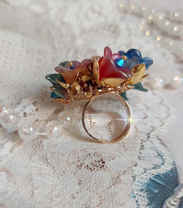 Bague Bel Air mont&eacute;e avec des fleurs lucites peintes &agrave; la main aux couleurs : Bleu, Rose, Rouge, Argent, Blanc avec feuilles ondul&eacute;es Vert Cuivr&eacute; &agrave; laquelle s&rsquo;ajoute perles Dor&eacute;es en Verre