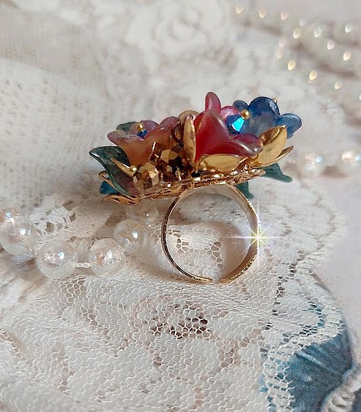 Bague Bel Air mont&eacute;e avec des fleurs lucites peintes &agrave; la main aux couleurs : Bleu, Rose, Rouge, Argent, Blanc avec feuilles ondul&eacute;es Vert Cuivr&eacute; &agrave; laquelle s&rsquo;ajoute perles Dor&eacute;es en Verre