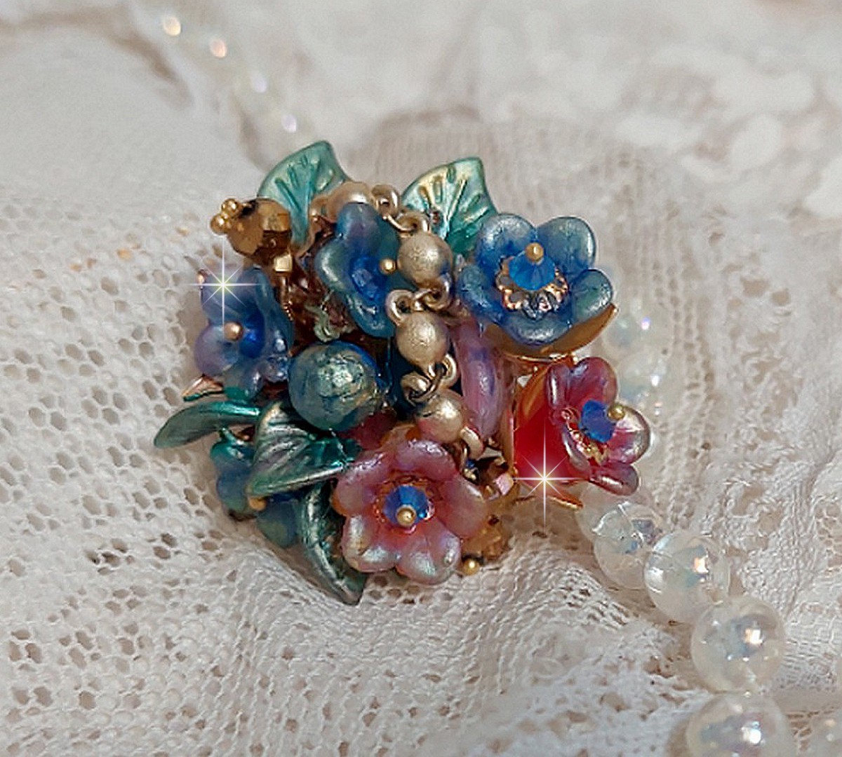 Bague Bel Air mont&eacute;e avec des fleurs lucites peintes &agrave; la main aux couleurs : Bleu, Rose, Rouge, Argent, Blanc avec feuilles ondul&eacute;es Vert Cuivr&eacute; &agrave; laquelle s&rsquo;ajoute perles Dor&eacute;es en Verre