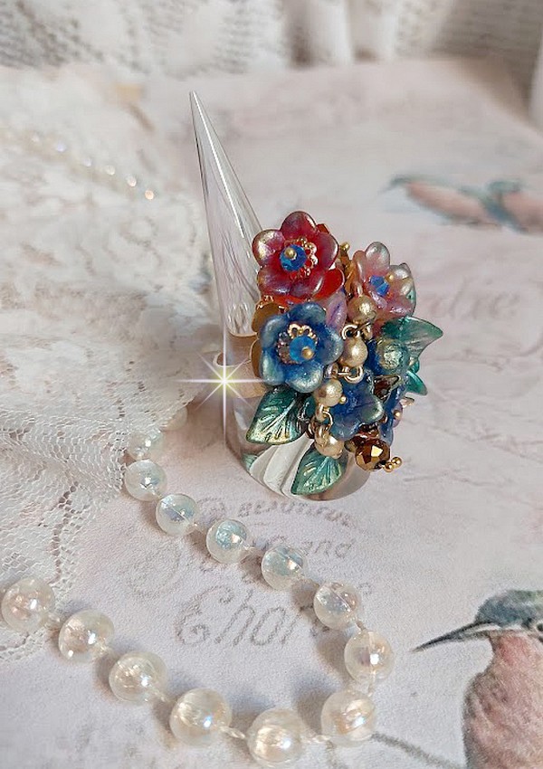 Bague Bel Air mont&eacute;e avec des fleurs lucites peintes &agrave; la main aux couleurs : Bleu, Rose, Rouge, Argent, Blanc avec feuilles ondul&eacute;es Vert Cuivr&eacute; &agrave; laquelle s&rsquo;ajoute perles Dor&eacute;es en Verre