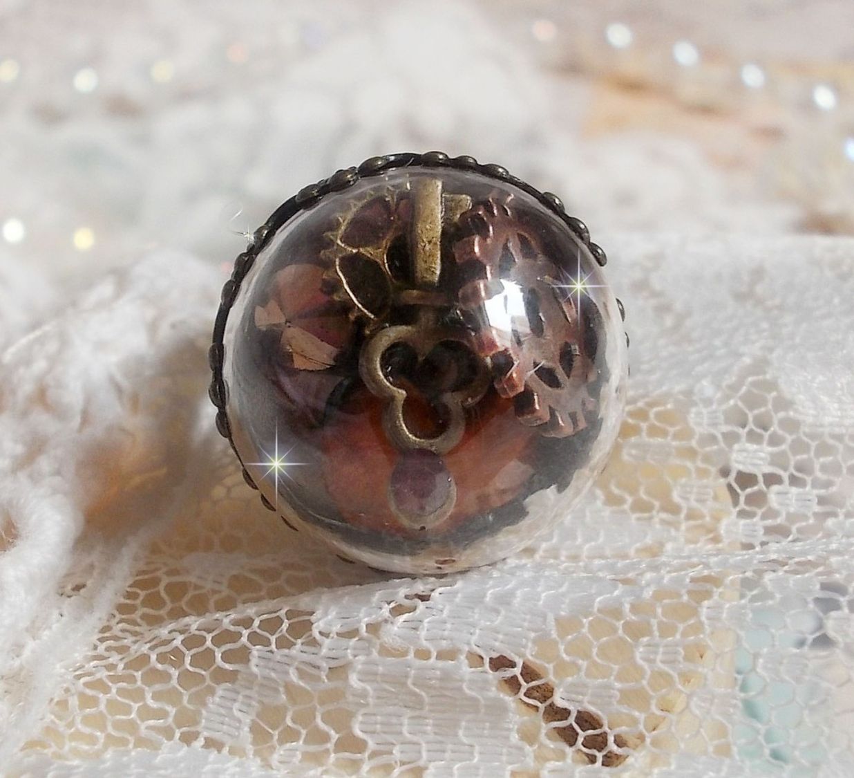 Bague Majest&eacute; Steampunk aux rouages m&eacute;caniques &agrave; laquelle s&rsquo;ajoute une cl&eacute; et un strass de couleur Siam et un strass en verre Dor&eacute;. Le tout est mis sous un globe en verre sur une bague r&eacute;glable laiton
