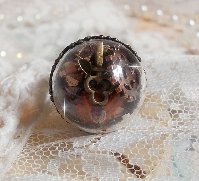 Bague Majest&eacute; Steampunk aux rouages m&eacute;caniques &agrave; laquelle s&rsquo;ajoute une cl&eacute; et un strass de couleur Siam et un strass en verre Dor&eacute;. Le tout est mis sous un globe en verre sur une bague r&eacute;glable laiton