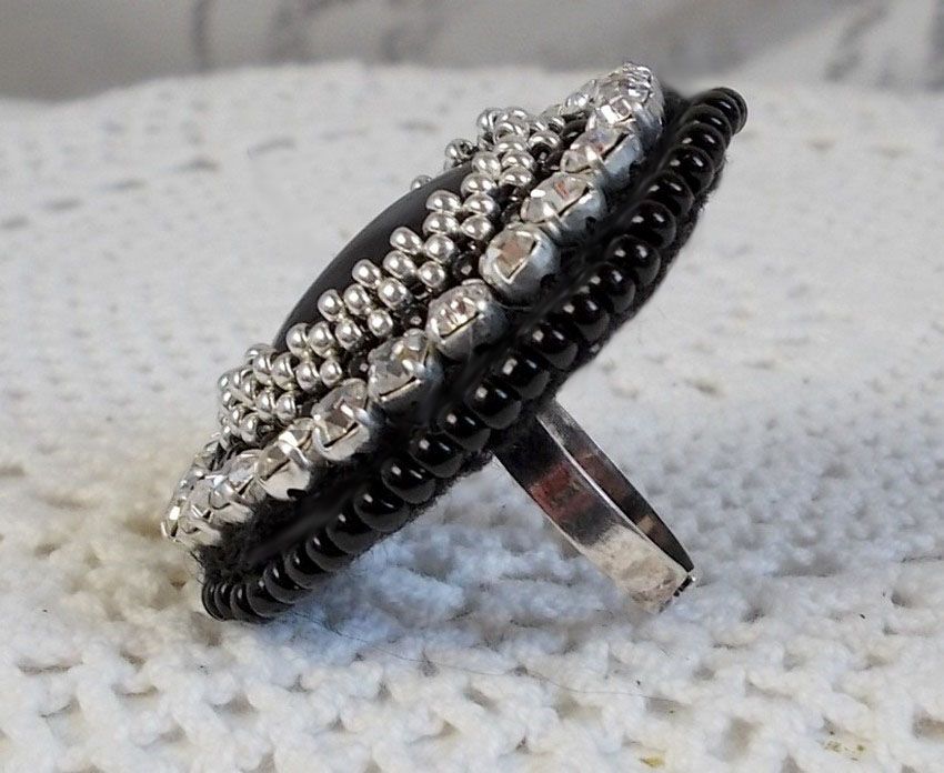 Bague Stone Noire avec un cabochon en Onyx orn&eacute; de chatons en Cristal &agrave; laquelle s&rsquo;ajoute des rocailles Argent&eacute;es qui compl&egrave;tent le Noir sur bague Nickel en Laiton et une feutrine. Un style R&eacute;tro