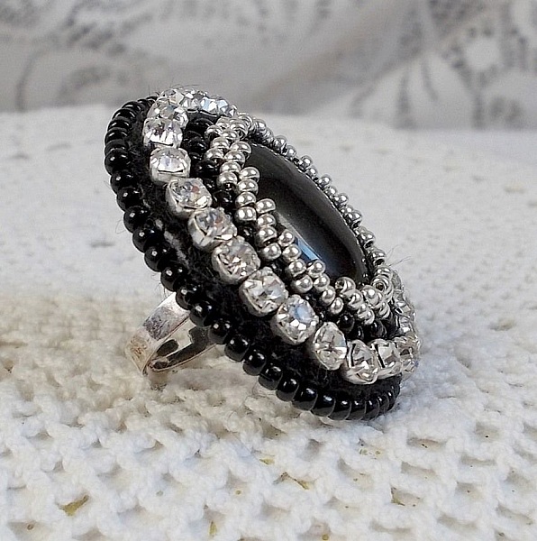 Bague Stone Noire avec un cabochon en Onyx orn&eacute; de chatons en Cristal &agrave; laquelle s&rsquo;ajoute des rocailles Argent&eacute;es qui compl&egrave;tent le Noir sur bague Nickel en Laiton et une feutrine. Un style R&eacute;tro