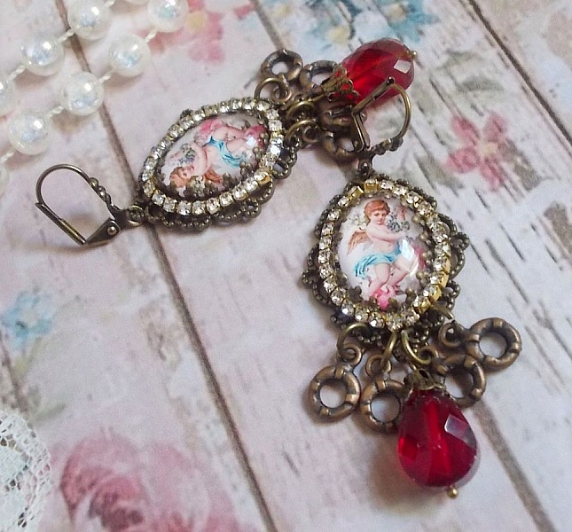 BO Petit Ange avec des cabochons d&rsquo;anges orn&eacute;s d&rsquo;une cha&icirc;ne strass Cristal, breloques, gouttes facett&eacute;es en verre de couleur Siam. Mont&eacute;s sur des pendentifs et des dormeuses en Laiton