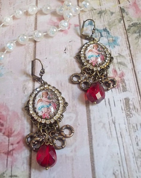 BO Petit Ange avec des cabochons d&rsquo;anges orn&eacute;s d&rsquo;une cha&icirc;ne strass Cristal, breloques, gouttes facett&eacute;es en verre de couleur Siam. Mont&eacute;s sur des pendentifs et des dormeuses en Laiton
