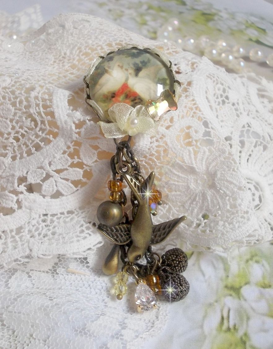Broche Colombes aux plumages blancs avec des breloques filigran&eacute;es gouttes et lisses, perles rondes et n&oelig;ud en cristal, ruban organza cr&egrave;me, pendentif plaqu&eacute; or avec un zircon sur une broche en m&eacute;tal