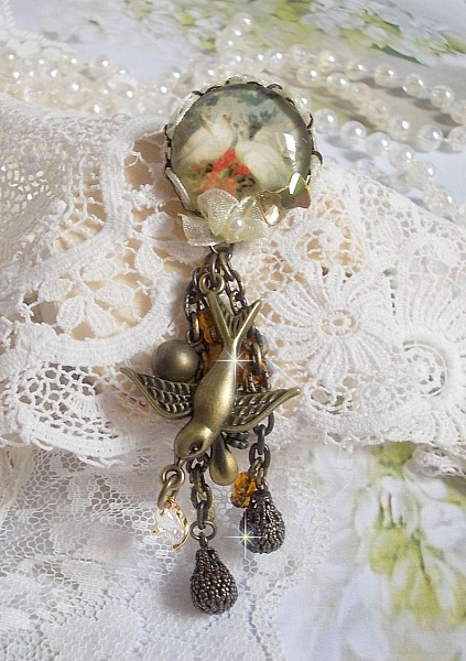 Broche Colombes aux plumages blancs avec des breloques filigran&eacute;es gouttes et lisses, perles rondes et n&oelig;ud en cristal, ruban organza cr&egrave;me, pendentif plaqu&eacute; or avec un zircon sur une broche en m&eacute;tal