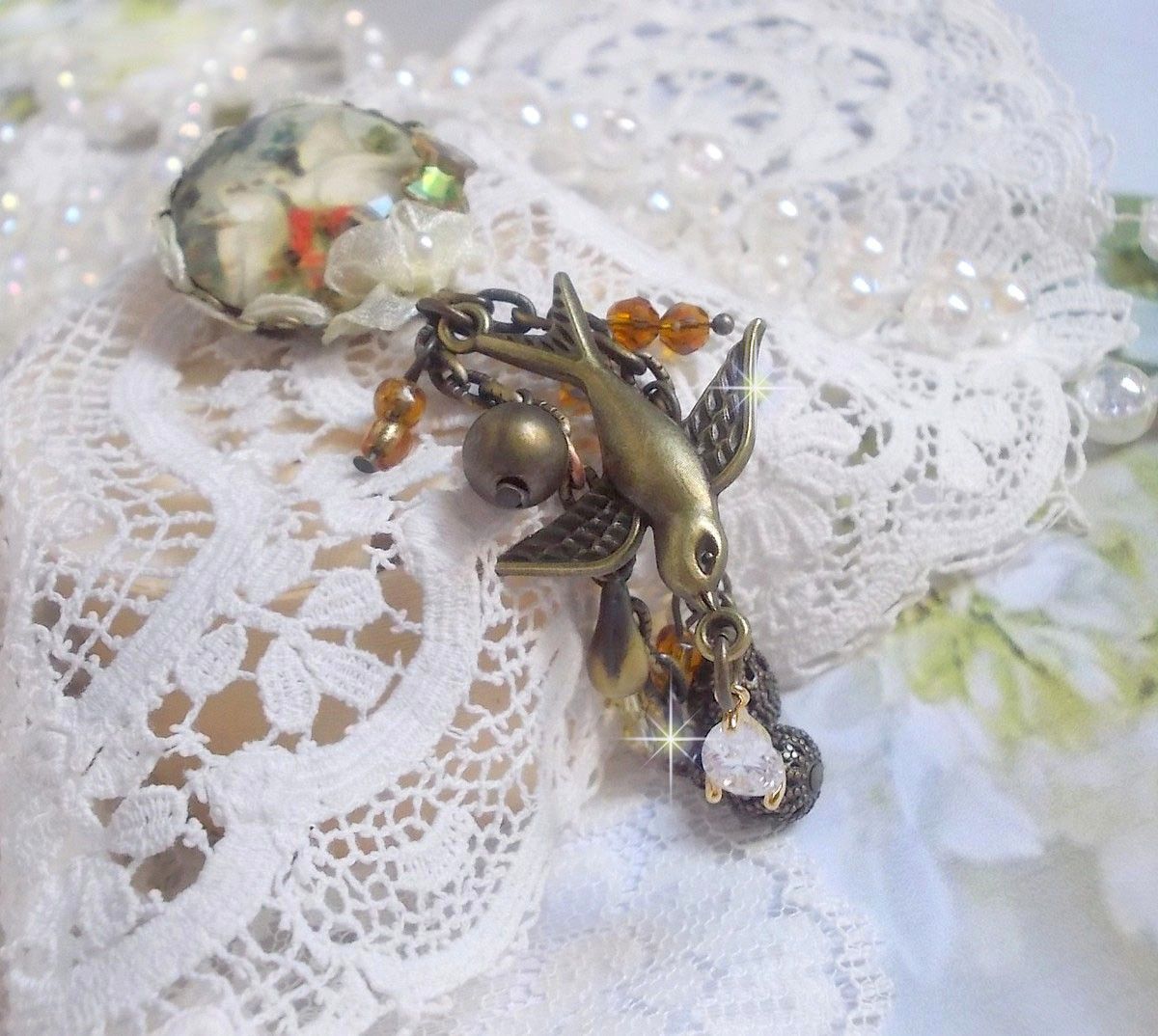 Broche Colombes aux plumages blancs avec des breloques filigran&eacute;es gouttes et lisses, perles rondes et n&oelig;ud en cristal, ruban organza cr&egrave;me, pendentif plaqu&eacute; or avec un zircon sur une broche en m&eacute;tal