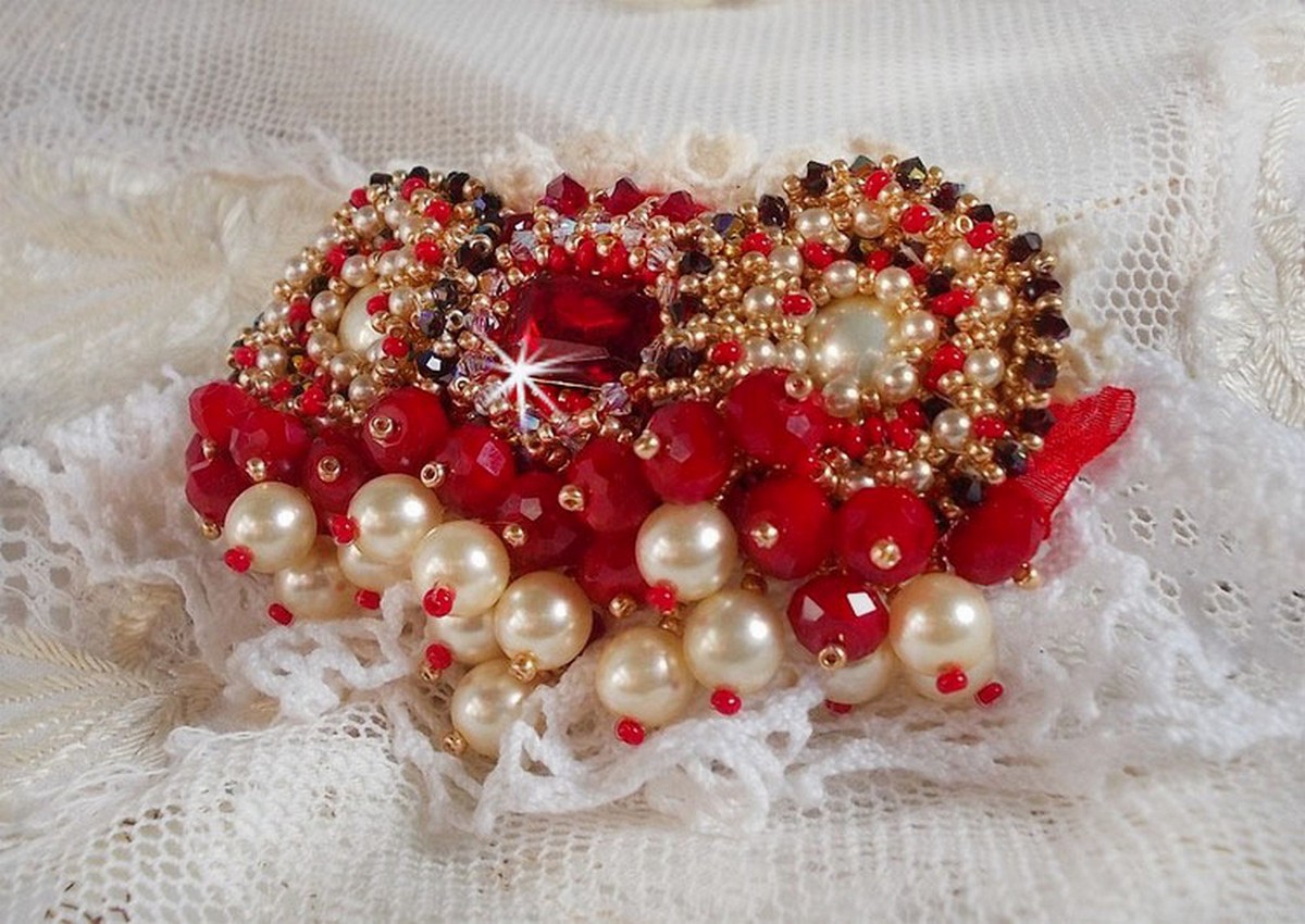 Broche Rubis brod&eacute;e avec trois cabochons Ivoire et Rouge &agrave; laquelle s&rsquo;ajoute des toupies aux couleurs Cristal, Burgundy et Garnet, des perles nacr&eacute;es et opaque. Mont&eacute; sur une dentelle et une broche