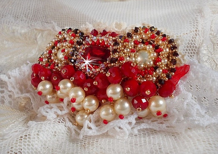 Broche Rubis brod&eacute;e avec trois cabochons Ivoire et Rouge &agrave; laquelle s&rsquo;ajoute des toupies aux couleurs Cristal, Burgundy et Garnet, des perles nacr&eacute;es et opaque. Mont&eacute; sur une dentelle et une broche