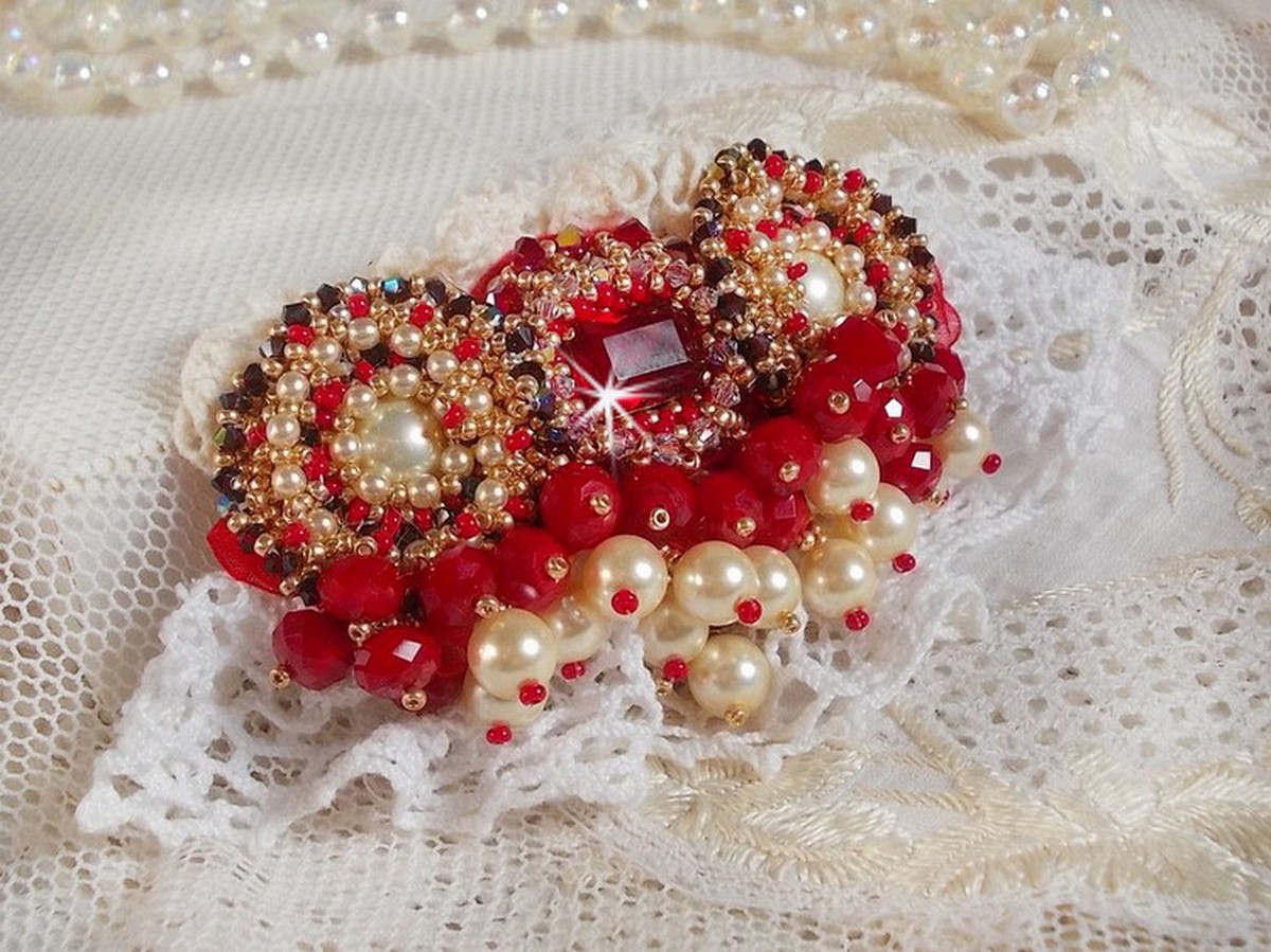Broche Rubis brod&eacute;e avec trois cabochons Ivoire et Rouge &agrave; laquelle s&rsquo;ajoute des toupies aux couleurs Cristal, Burgundy et Garnet, des perles nacr&eacute;es et opaque. Mont&eacute; sur une dentelle et une broche