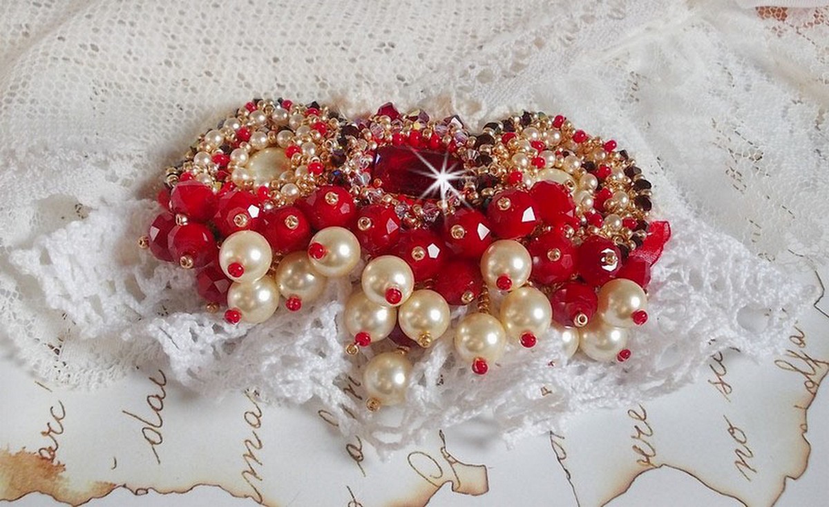Broche Rubis brod&eacute;e avec trois cabochons Ivoire et Rouge &agrave; laquelle s&rsquo;ajoute des toupies aux couleurs Cristal, Burgundy et Garnet, des perles nacr&eacute;es et opaque. Mont&eacute; sur une dentelle et une broche