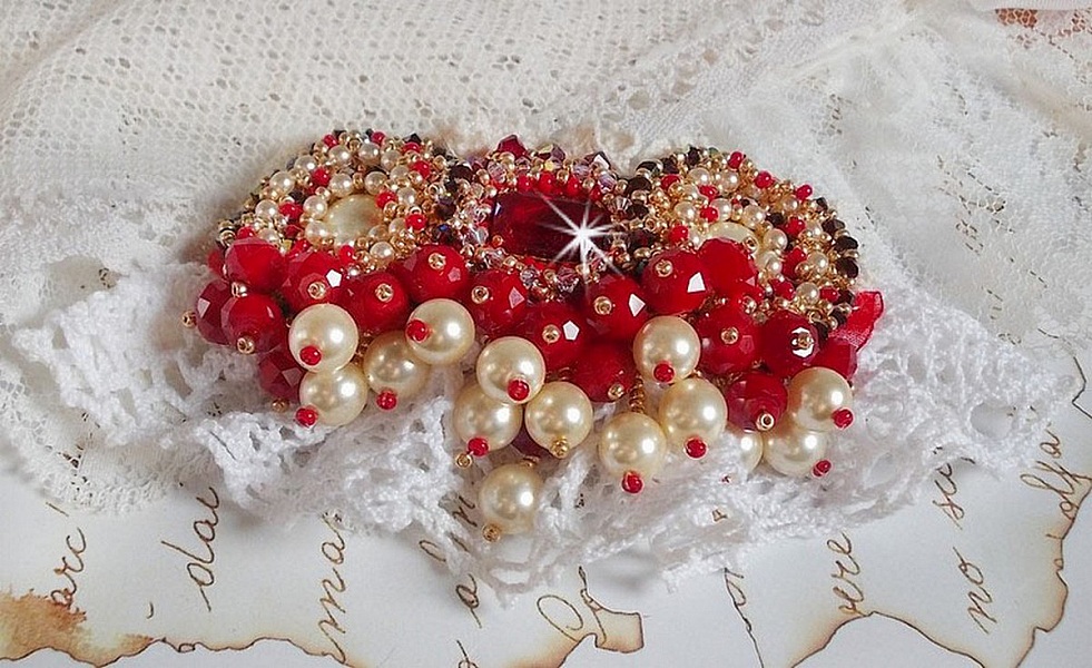 Broche Rubis brod&eacute;e avec trois cabochons Ivoire et Rouge &agrave; laquelle s&rsquo;ajoute des toupies aux couleurs Cristal, Burgundy et Garnet, des perles nacr&eacute;es et opaque. Mont&eacute; sur une dentelle et une broche