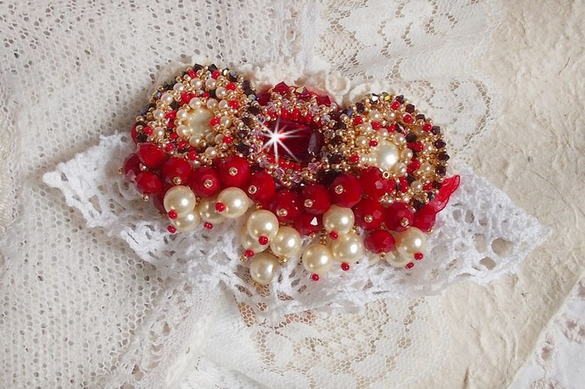 Broche Rubis brod&eacute;e avec trois cabochons Ivoire et Rouge &agrave; laquelle s&rsquo;ajoute des toupies aux couleurs Cristal, Burgundy et Garnet, des perles nacr&eacute;es et opaque. Mont&eacute; sur une dentelle et une broche