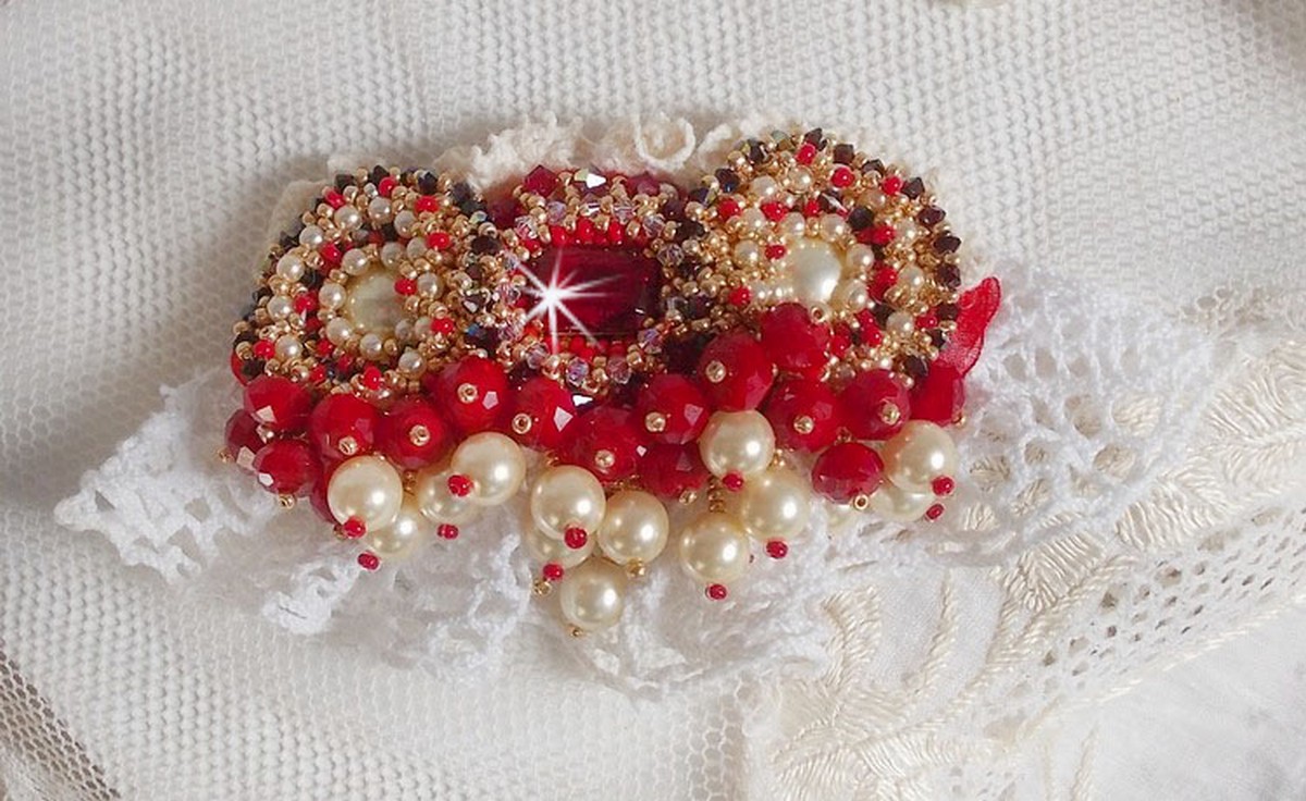 Broche Rubis brod&eacute;e avec trois cabochons Ivoire et Rouge &agrave; laquelle s&rsquo;ajoute des toupies aux couleurs Cristal, Burgundy et Garnet, des perles nacr&eacute;es et opaque. Mont&eacute; sur une dentelle et une broche