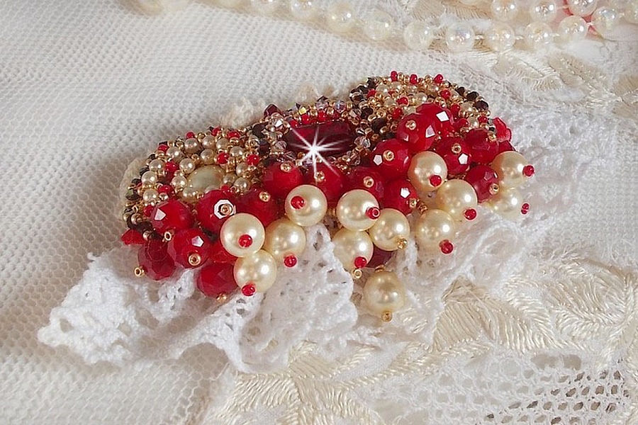 Broche Rubis brod&eacute;e avec trois cabochons Ivoire et Rouge &agrave; laquelle s&rsquo;ajoute des toupies aux couleurs Cristal, Burgundy et Garnet, des perles nacr&eacute;es et opaque. Mont&eacute; sur une dentelle et une broche
