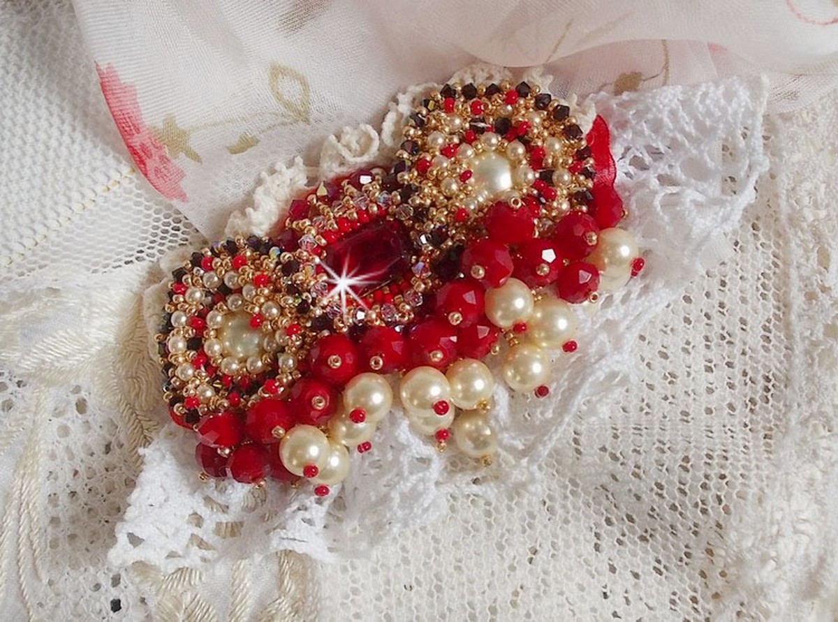 Broche Rubis brod&eacute;e avec trois cabochons Ivoire et Rouge &agrave; laquelle s&rsquo;ajoute des toupies aux couleurs Cristal, Burgundy et Garnet, des perles nacr&eacute;es et opaque. Mont&eacute; sur une dentelle et une broche