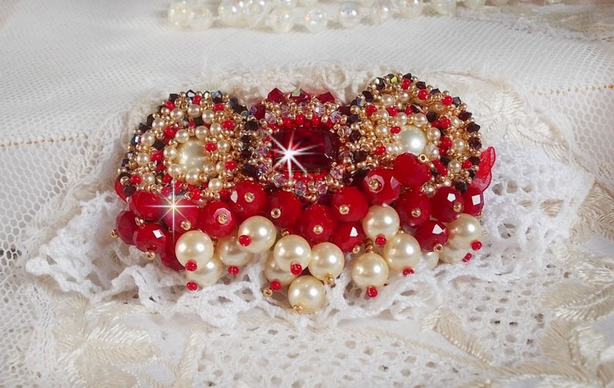 Broche Rubis brod&eacute;e avec trois cabochons Ivoire et Rouge &agrave; laquelle s&rsquo;ajoute des toupies aux couleurs Cristal, Burgundy et Garnet, des perles nacr&eacute;es et opaque. Mont&eacute; sur une dentelle et une broche