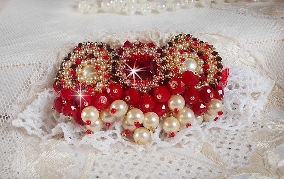 Broche Rubis brod&eacute;e avec trois cabochons Ivoire et Rouge &agrave; laquelle s&rsquo;ajoute des toupies aux couleurs Cristal, Burgundy et Garnet, des perles nacr&eacute;es et opaque. Mont&eacute; sur une dentelle et une broche