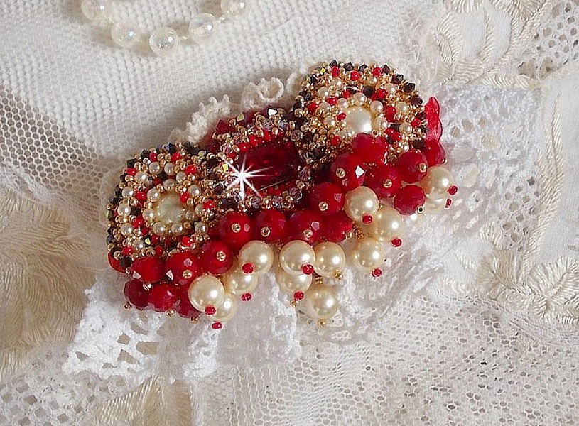 Broche Rubis brod&eacute;e avec trois cabochons Ivoire et Rouge &agrave; laquelle s&rsquo;ajoute des toupies aux couleurs Cristal, Burgundy et Garnet, des perles nacr&eacute;es et opaque. Mont&eacute; sur une dentelle et une broche