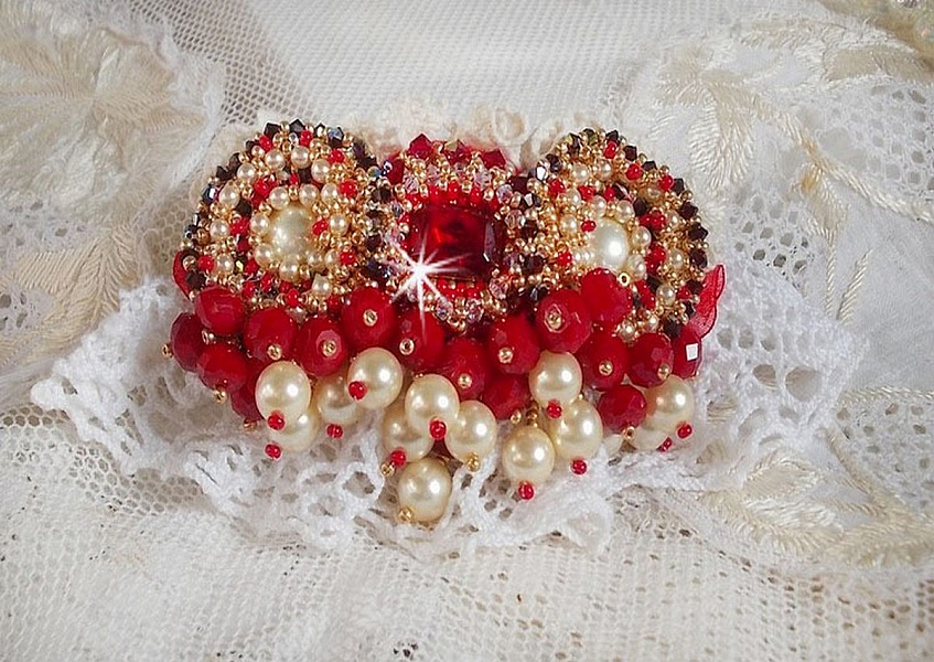 Broche Rubis brod&eacute;e avec trois cabochons Ivoire et Rouge &agrave; laquelle s&rsquo;ajoute des toupies aux couleurs Cristal, Burgundy et Garnet, des perles nacr&eacute;es et opaque. Mont&eacute; sur une dentelle et une broche