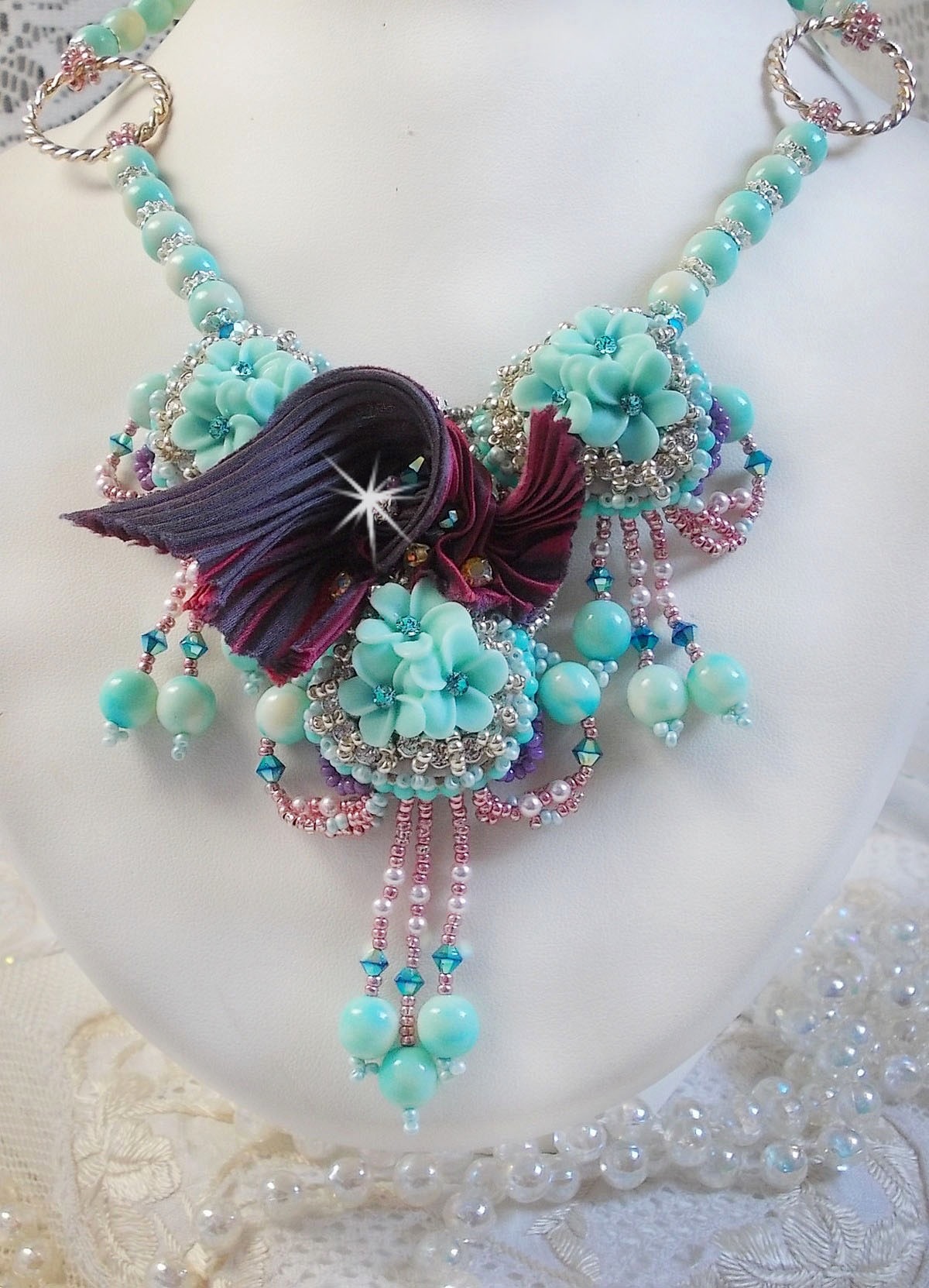 Collier Blue Flowers brod&eacute; avec un ruban de soie framboise/truffe &agrave; laquelle s&rsquo;ajoute : cabochons, perles nacr&eacute;es Rosaline et Ivoire, toupies Jet, anneaux ovales, perles marbr&eacute;es Bleu Ciel et Blanc