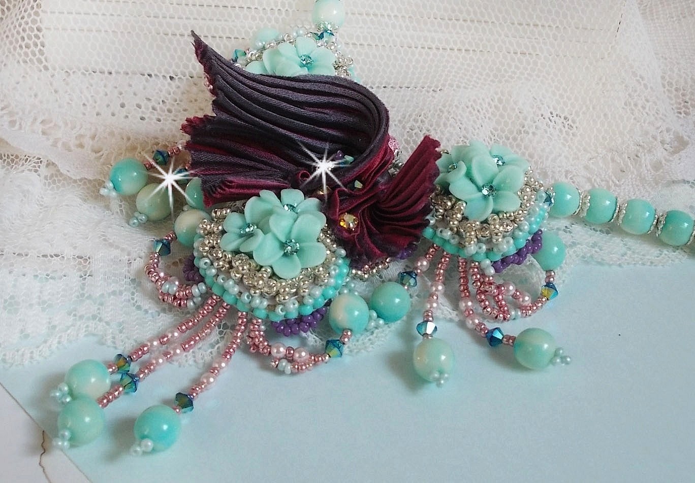 Collier Blue Flowers brod&eacute; avec un ruban de soie framboise/truffe &agrave; laquelle s&rsquo;ajoute : cabochons, perles nacr&eacute;es Rosaline et Ivoire, toupies Jet, anneaux ovales, perles marbr&eacute;es Bleu Ciel et Blanc