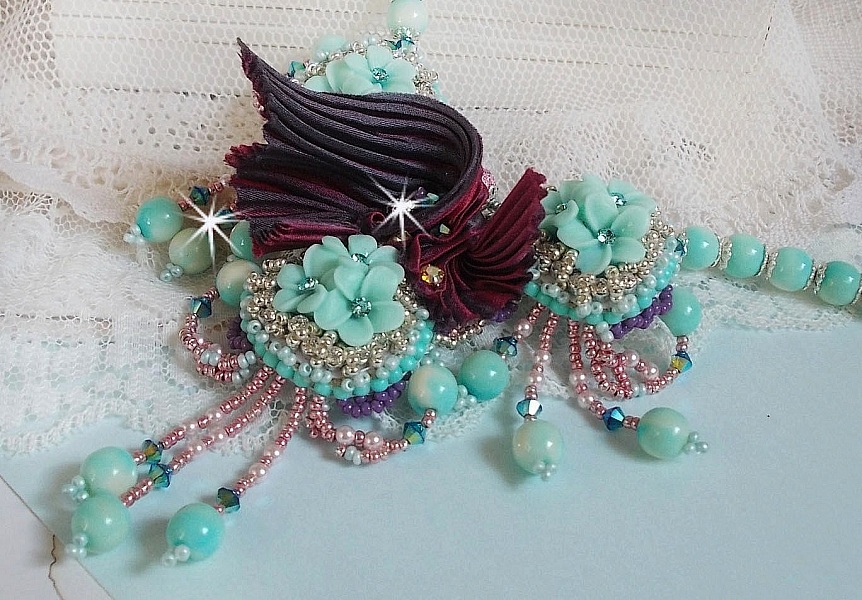 Collier Blue Flowers brod&eacute; avec un ruban de soie framboise/truffe &agrave; laquelle s&rsquo;ajoute : cabochons, perles nacr&eacute;es Rosaline et Ivoire, toupies Jet, anneaux ovales, perles marbr&eacute;es Bleu Ciel et Blanc