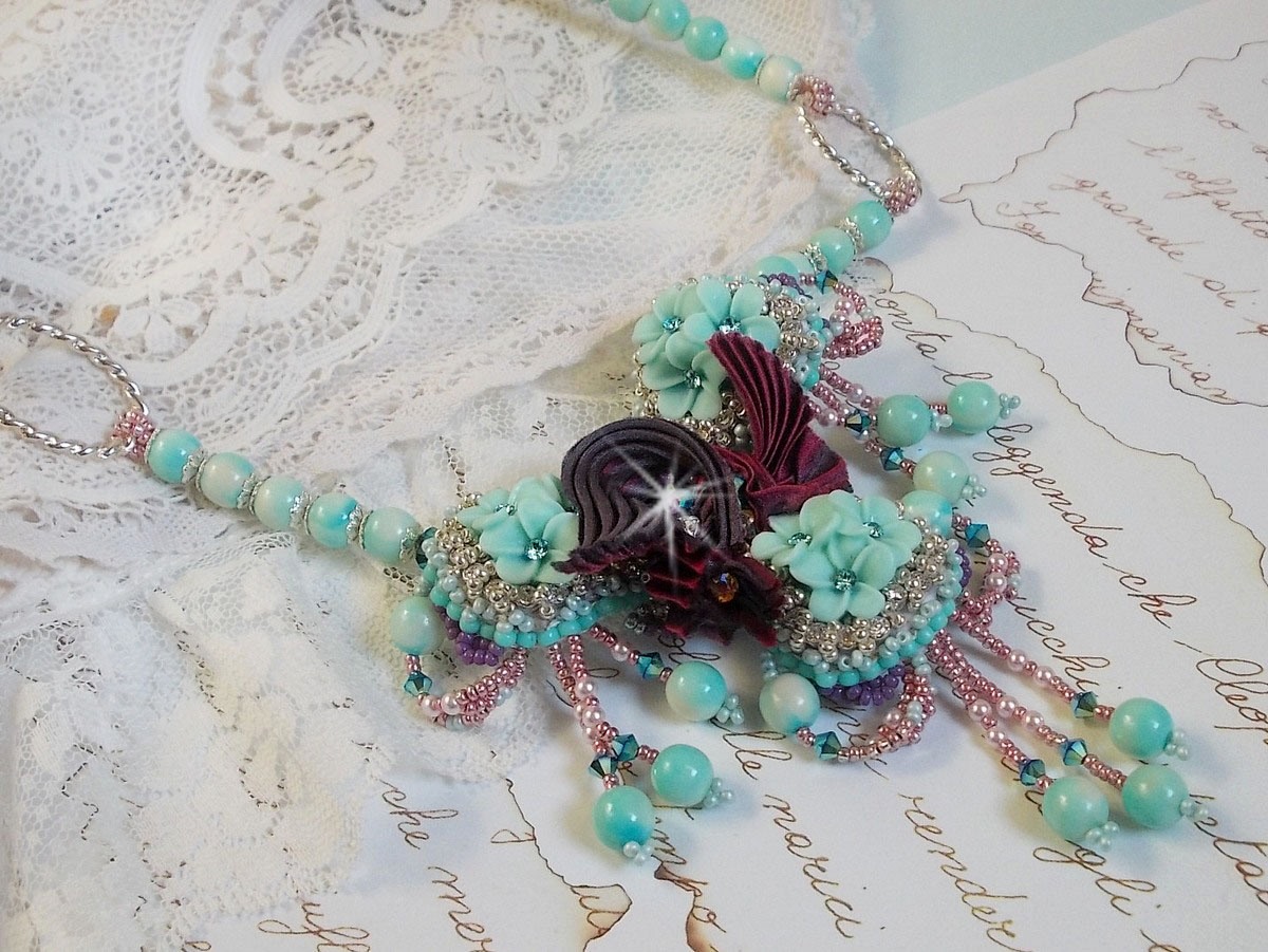 Collier Blue Flowers brod&eacute; avec un ruban de soie framboise/truffe &agrave; laquelle s&rsquo;ajoute : cabochons, perles nacr&eacute;es Rosaline et Ivoire, toupies Jet, anneaux ovales, perles marbr&eacute;es Bleu Ciel et Blanc