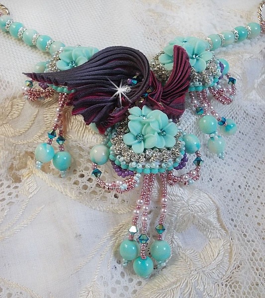 Collier Blue Flowers brod&eacute; avec un ruban de soie framboise/truffe &agrave; laquelle s&rsquo;ajoute : cabochons, perles nacr&eacute;es Rosaline et Ivoire, toupies Jet, anneaux ovales, perles marbr&eacute;es Bleu Ciel et Blanc
