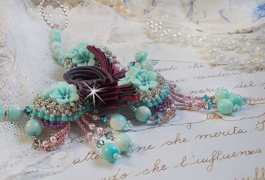 Collier Blue Flowers brod&eacute; avec un ruban de soie framboise/truffe &agrave; laquelle s&rsquo;ajoute : cabochons, perles nacr&eacute;es Rosaline et Ivoire, toupies Jet, anneaux ovales, perles marbr&eacute;es Bleu Ciel et Blanc