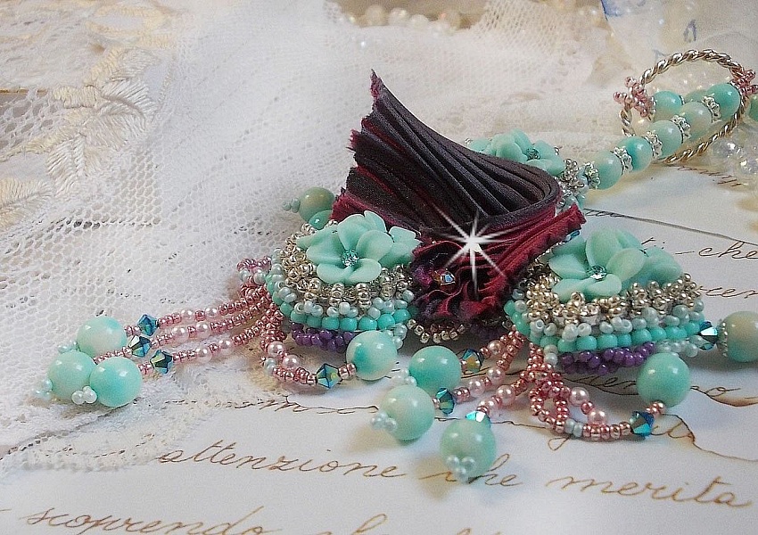 Collier Blue Flowers brod&eacute; avec un ruban de soie framboise/truffe &agrave; laquelle s&rsquo;ajoute : cabochons, perles nacr&eacute;es Rosaline et Ivoire, toupies Jet, anneaux ovales, perles marbr&eacute;es Bleu Ciel et Blanc