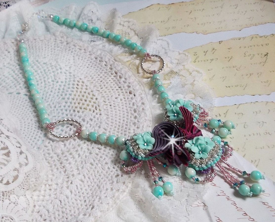 Collier Blue Flowers brod&eacute; avec un ruban de soie framboise/truffe &agrave; laquelle s&rsquo;ajoute : cabochons, perles nacr&eacute;es Rosaline et Ivoire, toupies Jet, anneaux ovales, perles marbr&eacute;es Bleu Ciel et Blanc