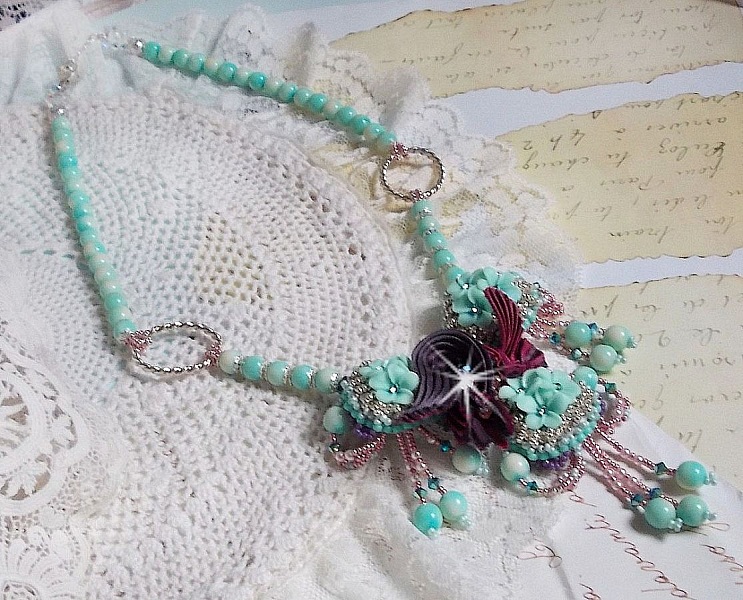 Collier Blue Flowers brod&eacute; avec un ruban de soie framboise/truffe &agrave; laquelle s&rsquo;ajoute : cabochons, perles nacr&eacute;es Rosaline et Ivoire, toupies Jet, anneaux ovales, perles marbr&eacute;es Bleu Ciel et Blanc