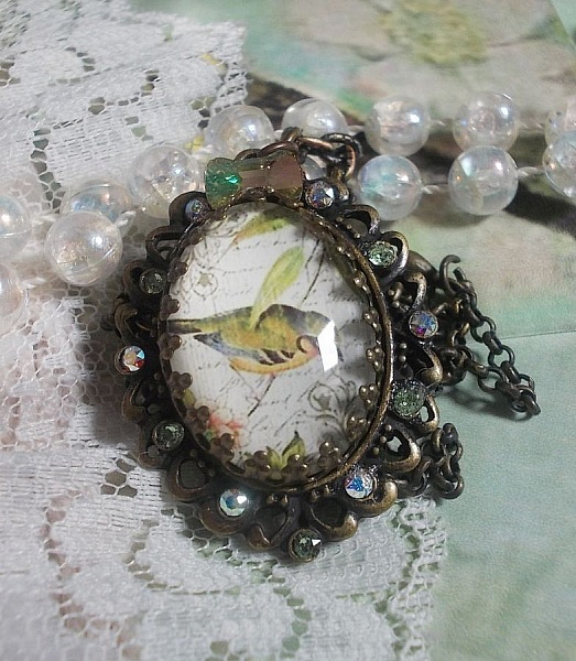 Collier Oiseau des Jardins s&rsquo;inspire d&rsquo;un oiseau dans la nature. Sur une base d&rsquo;un pendentif, ce motif est orn&eacute; de cristaux : n&oelig;ud et cabochons aux nuances cristal et P&eacute;ridot. Mont&eacute; sur cha&icirc;ne Laiton