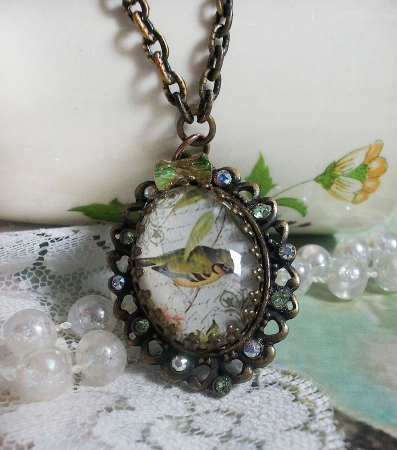 Collier Oiseau des Jardins s&rsquo;inspire d&rsquo;un oiseau dans la nature. Sur une base d&rsquo;un pendentif, ce motif est orn&eacute; de cristaux : n&oelig;ud et cabochons aux nuances cristal et P&eacute;ridot. Mont&eacute; sur cha&icirc;ne Laiton