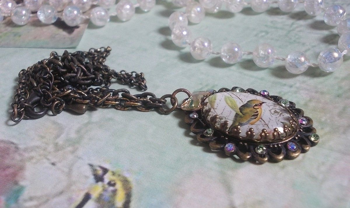 Collier Oiseau des Jardins s&rsquo;inspire d&rsquo;un oiseau dans la nature. Sur une base d&rsquo;un pendentif, ce motif est orn&eacute; de cristaux : n&oelig;ud et cabochons aux nuances cristal et P&eacute;ridot. Mont&eacute; sur cha&icirc;ne Laiton
