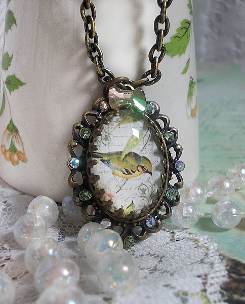 Collier Oiseau des Jardins s&rsquo;inspire d&rsquo;un oiseau dans la nature. Sur une base d&rsquo;un pendentif, ce motif est orn&eacute; de cristaux : n&oelig;ud et cabochons aux nuances cristal et P&eacute;ridot. Mont&eacute; sur cha&icirc;ne Laiton