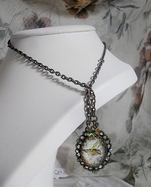 Collier Oiseau des Jardins s&rsquo;inspire d&rsquo;un oiseau dans la nature. Sur une base d&rsquo;un pendentif, ce motif est orn&eacute; de cristaux : n&oelig;ud et cabochons aux nuances cristal et P&eacute;ridot. Mont&eacute; sur cha&icirc;ne Laiton