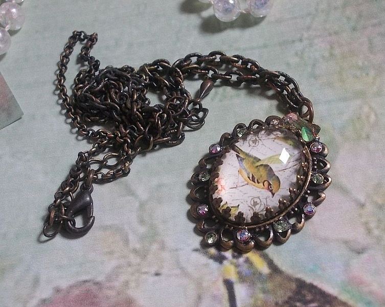 Collier Oiseau des Jardins s&rsquo;inspire d&rsquo;un oiseau dans la nature. Sur une base d&rsquo;un pendentif, ce motif est orn&eacute; de cristaux : n&oelig;ud et cabochons aux nuances cristal et P&eacute;ridot. Mont&eacute; sur cha&icirc;ne Laiton