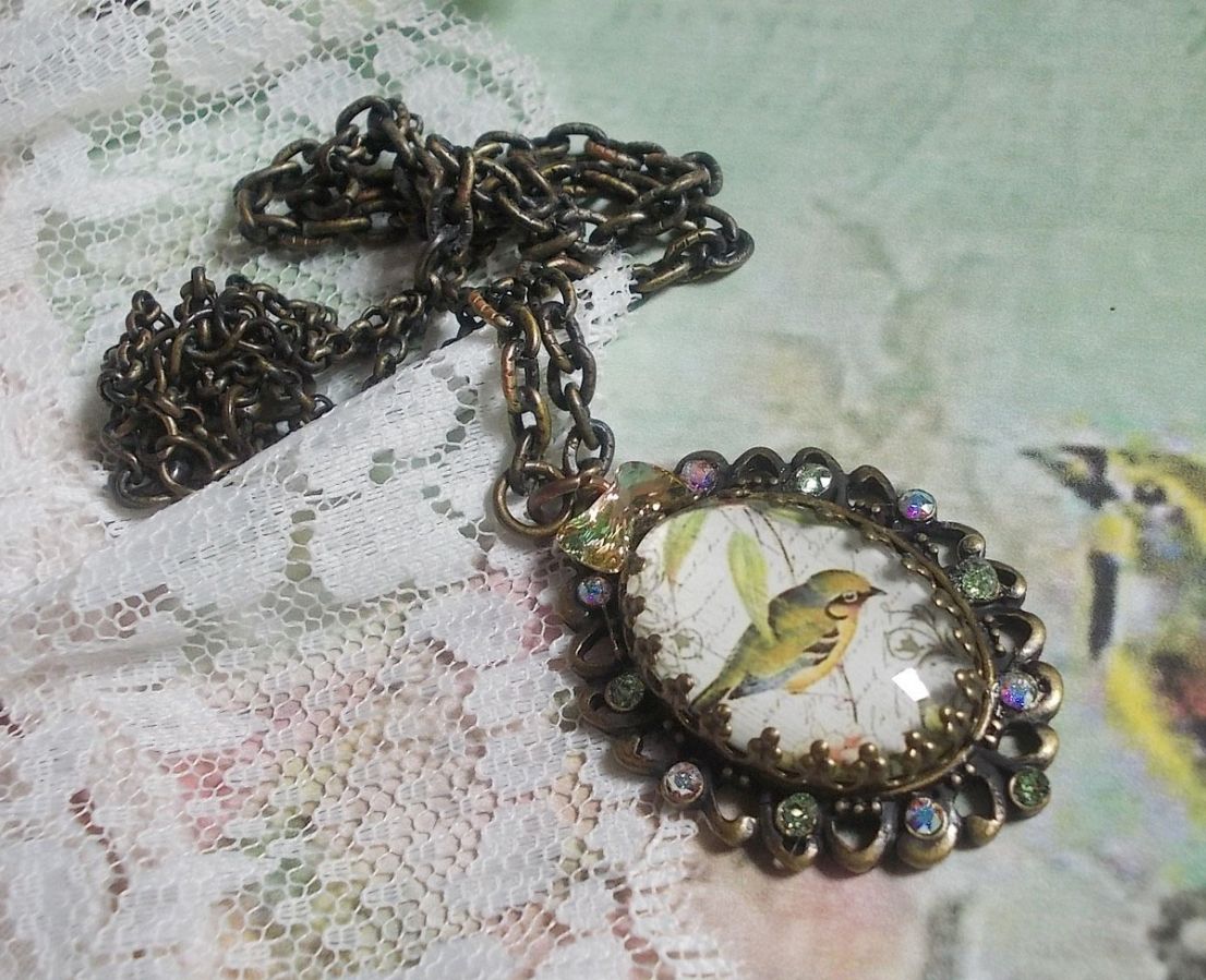 Collier Oiseau des Jardins s&rsquo;inspire d&rsquo;un oiseau dans la nature. Sur une base d&rsquo;un pendentif, ce motif est orn&eacute; de cristaux : n&oelig;ud et cabochons aux nuances cristal et P&eacute;ridot. Mont&eacute; sur cha&icirc;ne Laiton
