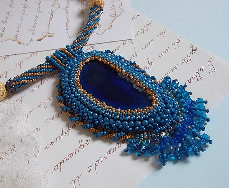 Pendentif Corsica avec un cabochon : une Agate Bleue brod&eacute;e avec des perles nacr&eacute;es Bleu Lapis en cristal &agrave; laquelle s&rsquo;ajoute des rocailles saphir et travertin entrelac&eacute;es pour former une dentelle