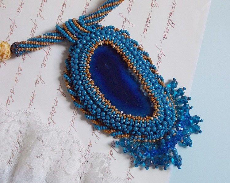 Pendentif Corsica avec un cabochon : une Agate Bleue brod&eacute;e avec des perles nacr&eacute;es Bleu Lapis en cristal &agrave; laquelle s&rsquo;ajoute des rocailles saphir et travertin entrelac&eacute;es pour former une dentelle