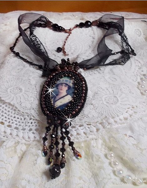 Pendentif Midnight Daisy avec un cabochon en bois repr&eacute;sentant une femme en chapeau Bleu, brod&eacute; avec des fleurs lucites, coupelles et facettes. Le tout est fini avec une dentelle Noire et un ruban